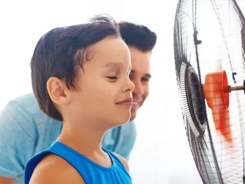 A young person stands with eyes closed in front of a pedestal fan, with someone smiling behind them.