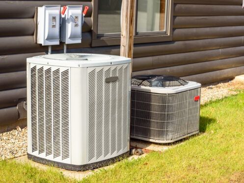 Two central air conditioning units sit on a concrete pad against the exterior wall of a log home.
