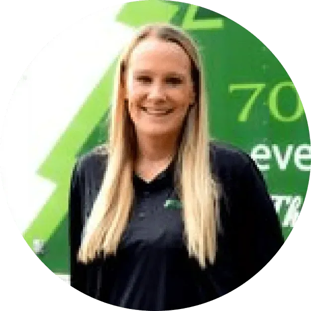 A person with long blonde hair smiling, wearing a dark polo shirt against a green background with partial text.