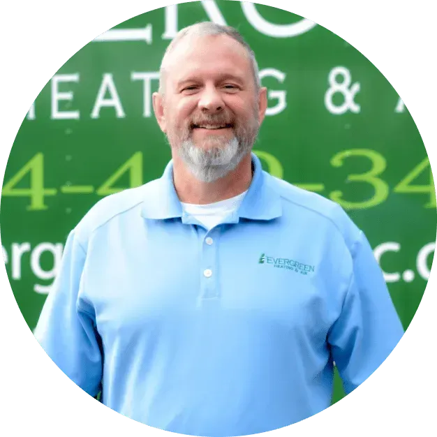 A smiling person wearing a light blue collared shirt stands in front of a green Evergreen Heating and Air service truck.