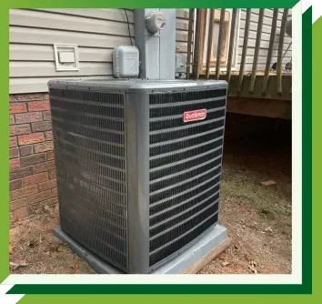 A grey Goodman brand outdoor HVAC condenser unit sits on a concrete pad next to a house with brick and siding walls.