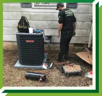 A technician services an outdoor Goodman air conditioning unit near the side of a house, with tools laid out on the ground.