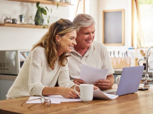 Two people sit at a kitchen counter reviewing documents on a laptop and smiling.