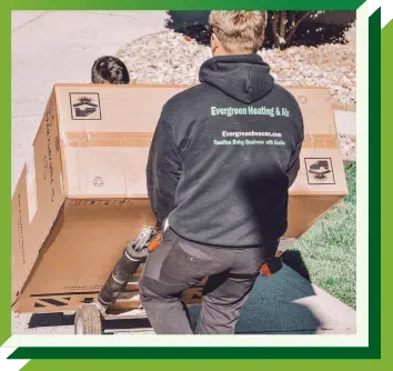 A worker in an Evergreen Heating & Air hoodie uses a dolly to move a large cardboard shipping box outdoors.