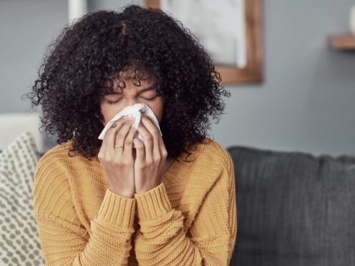 A person with curly hair wearing a yellow sweater blows their nose into a tissue while sitting on a couch.