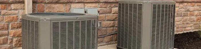 Two gray air conditioning units sitting on a concrete pad against a stone brick wall.