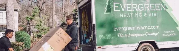 Two technicians carry a large box from an Evergreen Heating & Air service truck parked in a residential neighborhood.