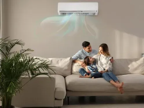 A family relaxes on a beige sofa in a room with a wall-mounted air conditioner blowing cool air.