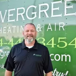 A person wearing a black polo shirt stands in front of a green Evergreen Heating & Air service truck.