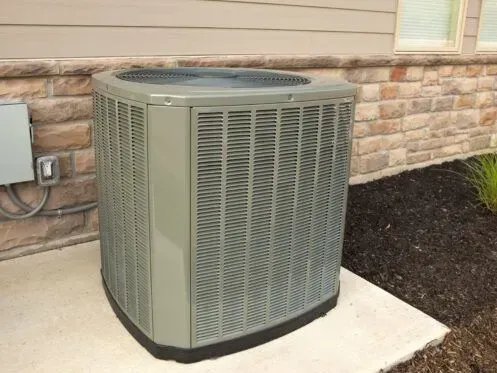 An outdoor gray air conditioning condenser unit placed on a concrete pad against a stone exterior wall.