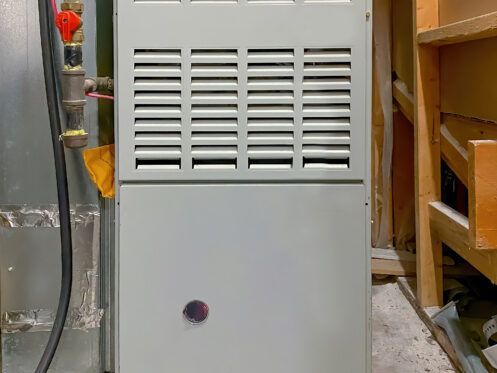A gray gas furnace with a red gas shut-off valve, positioned in an unfinished basement area with wooden framing.