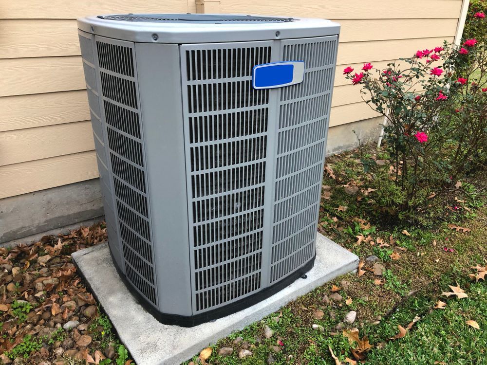 A gray residential air conditioning unit sits on a concrete slab in a yard next to a house with beige siding and roses.