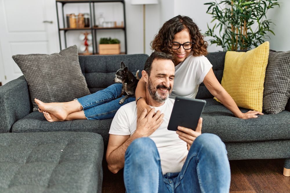 A couple sits together on a grey sofa in a living room, looking at a tablet with a small dog resting beside them.
