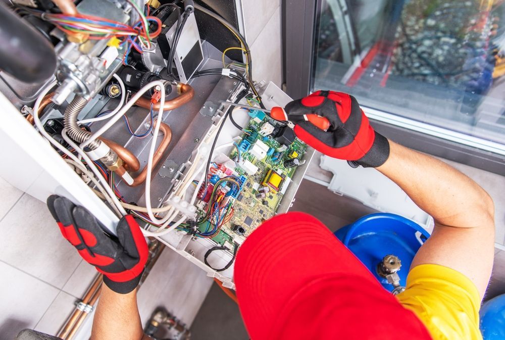 A technician in a red cap and gloves repairs an open wall-mounted boiler, focusing on the internal circuit board.