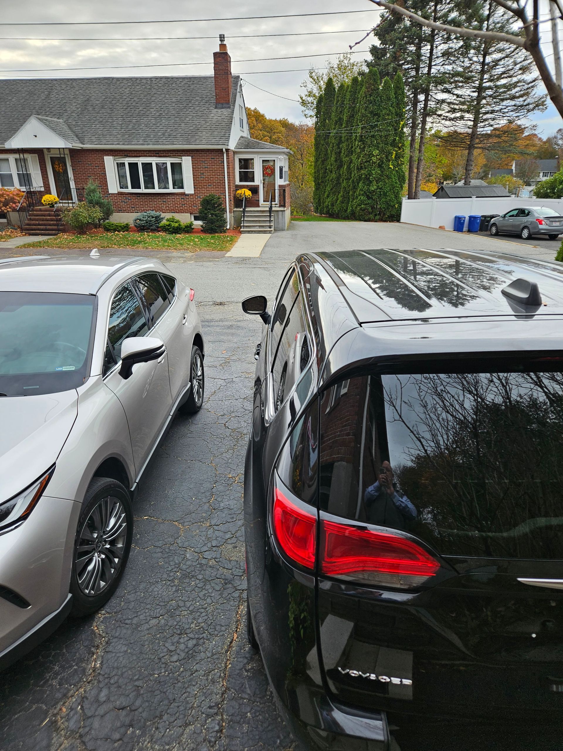 Parked cars in a driveway facing a brick house with a chimney and trees in the background.