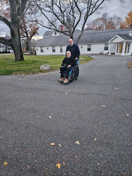 Two people on a wheelchair on a gravel driveway beside a white house and trees in autumn