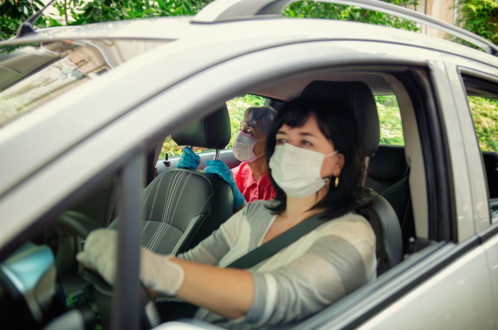 Masked driver in a white car at a drive-through window, with another passenger visible in back.