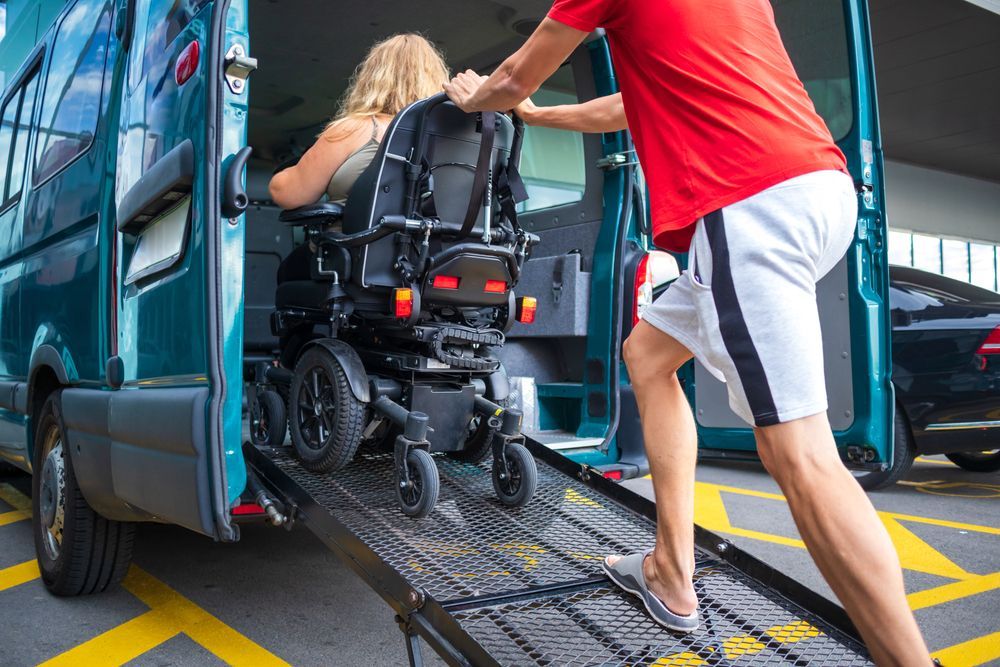 Person in wheelchair being pushed up a ramp into a blue van by another person in red shirt