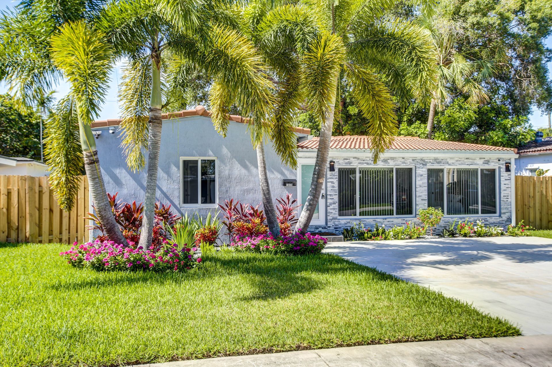 Single-story white house with a green lawn, palm trees, and colorful flowers beside a driveway