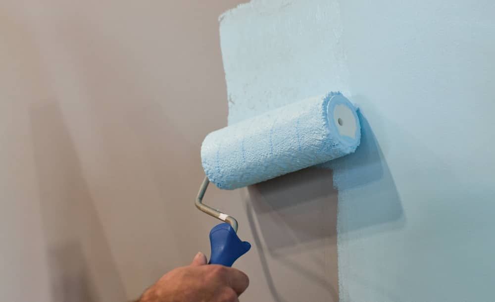 Hand using a blue paint roller to apply light blue paint to a white wall.