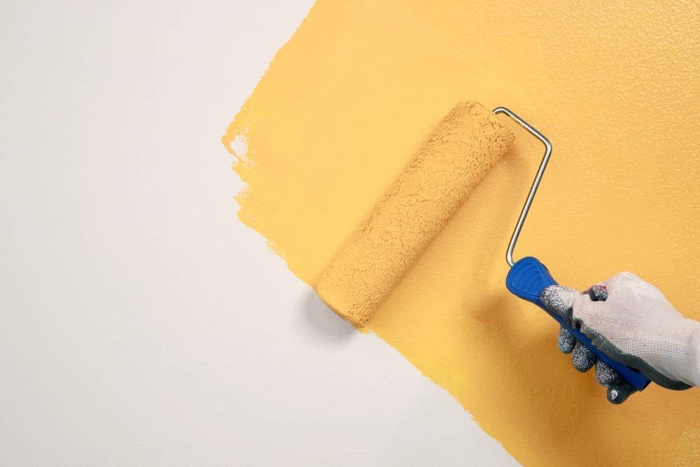 Hand in blue glove painting a wall yellow with a paint roller