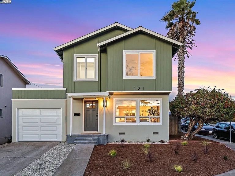 Two-story house with green siding, a garage, and a palm tree in the front yard at sunset.