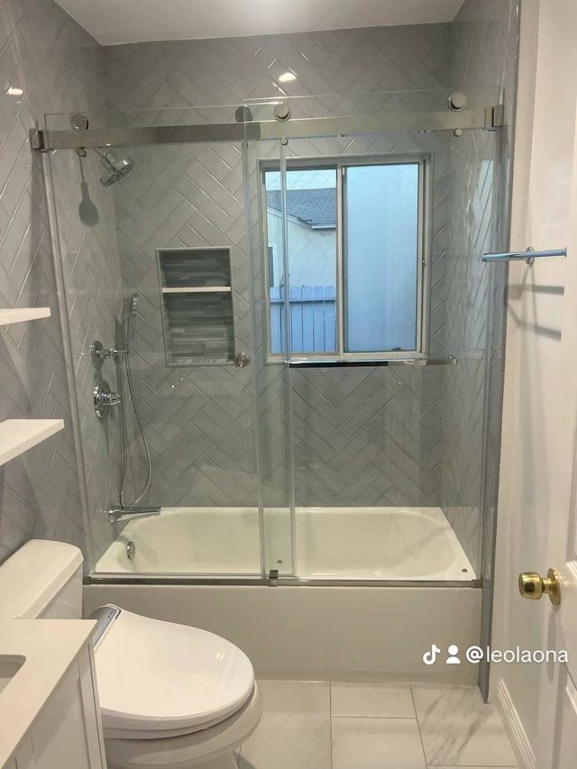Bathroom with gray herringbone tile, glass shower doors, white tub, and toilet.