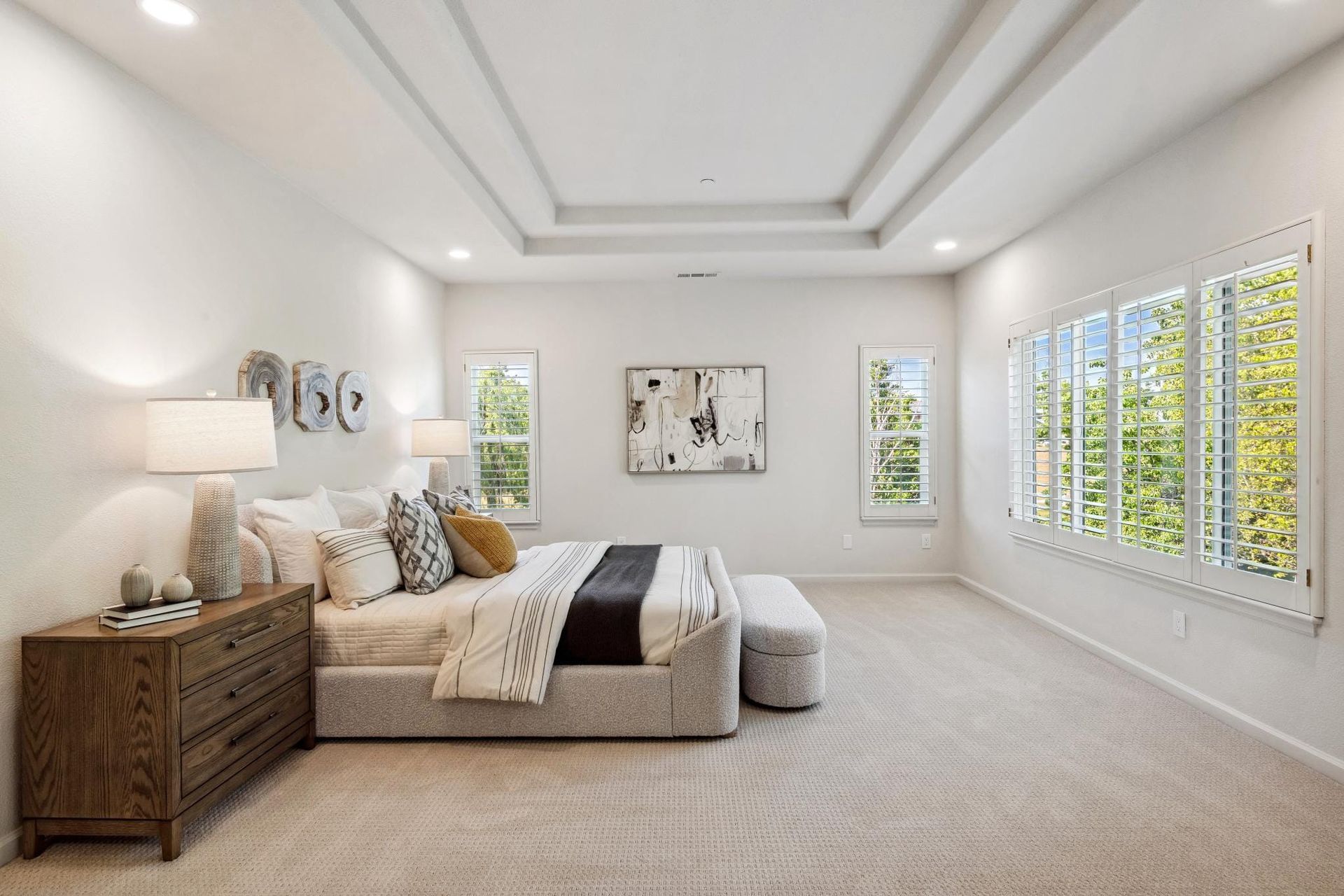 Spacious bedroom with light walls, beige carpet, king bed, wooden dresser, artwork, and large windows with shutters.