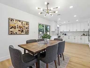 Dining room with wooden table, six gray chairs, and modern light fixture. White walls and kitchen.