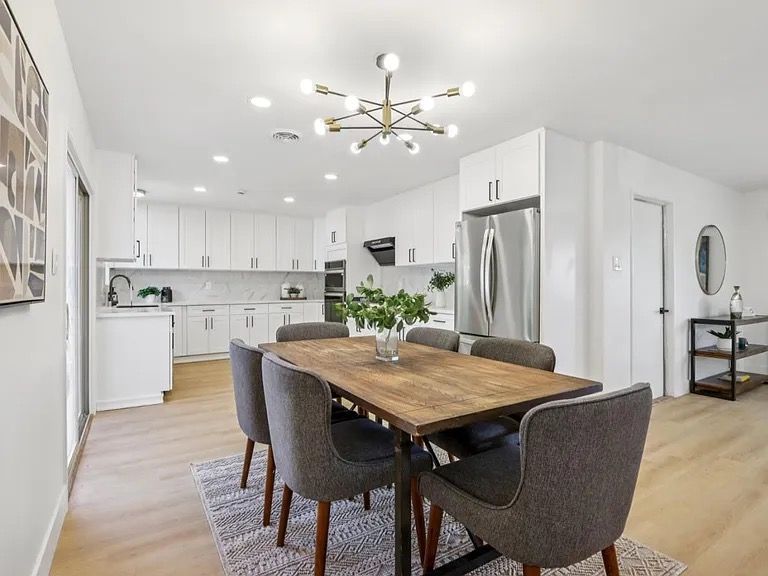 Dining room with wooden table, gray chairs, and modern light fixture. Open to white kitchen.