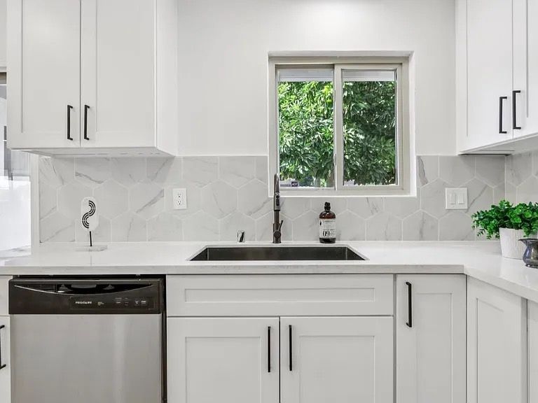 White kitchen with a black sink and faucet, stainless steel dishwasher, and a window with a view of greenery.