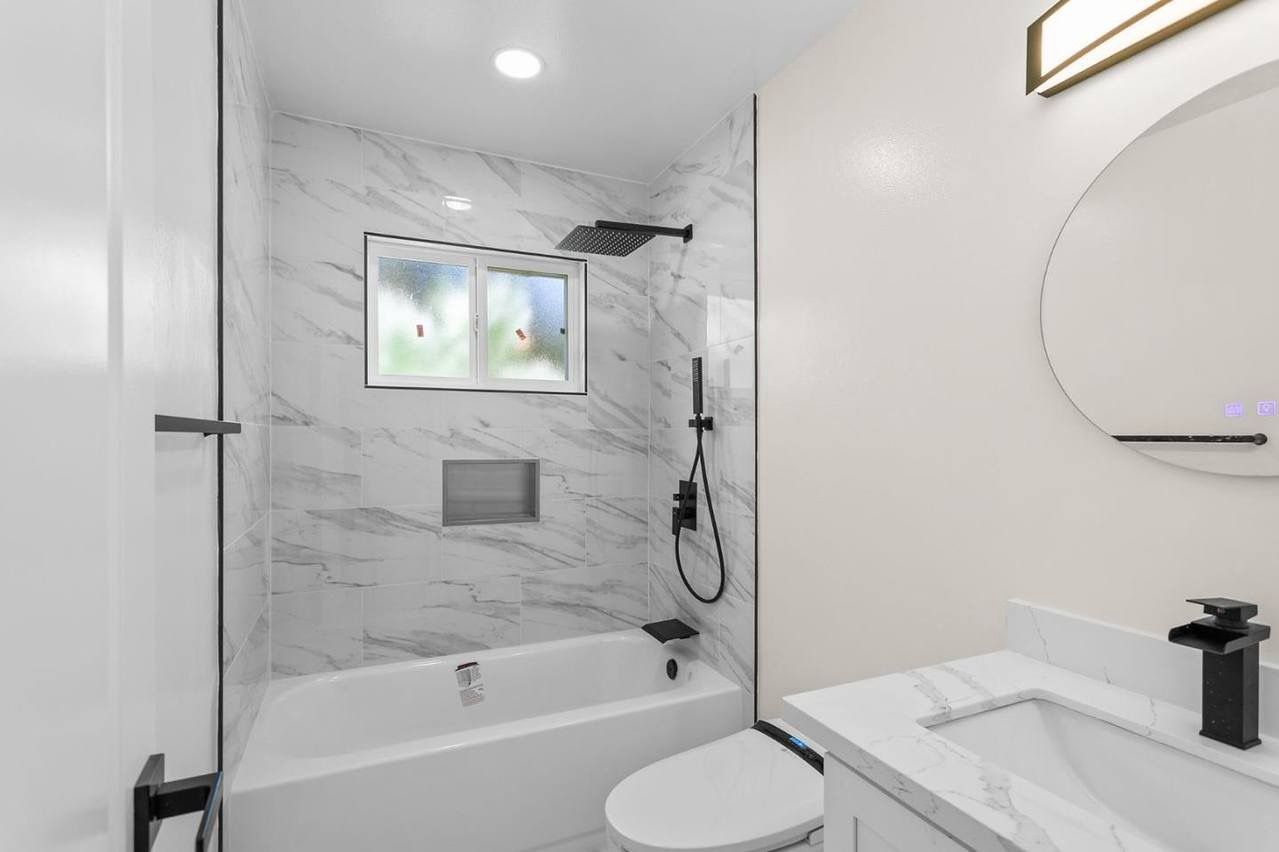 Modern white bathroom with marble shower, tub, toilet, vanity, and a round mirror. Black fixtures.