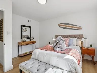 Bedroom with bed, bench, side table, mirror, surfboard decoration, and wood flooring.