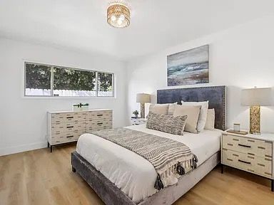 Bedroom with white walls, wood floors, bed with blue headboard, dressers, artwork, and natural light.