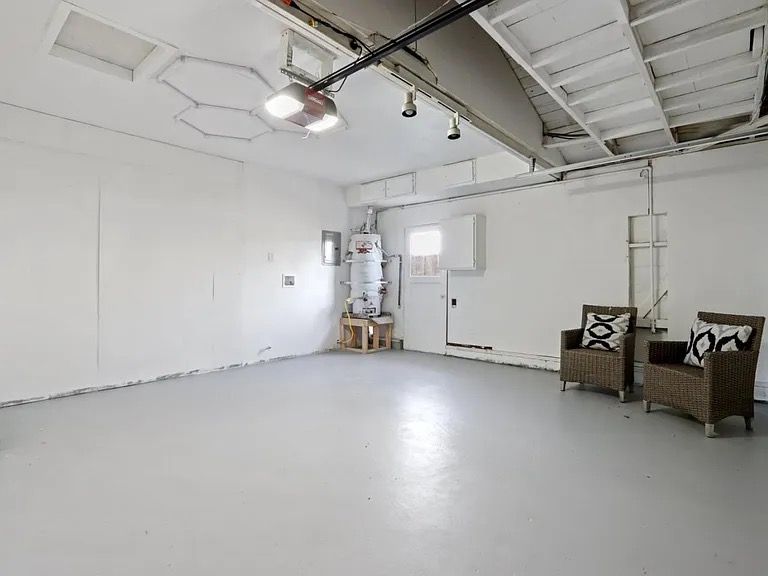 Empty, white-painted garage with light gray floor. Two wicker chairs sit near the right wall. Ceiling lights.
