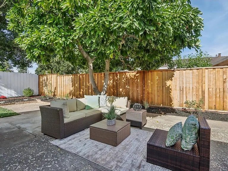 Patio with brown wicker furniture, rug, and wooden fence, under a large tree.