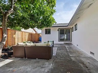 Backyard patio with brown wicker furniture under a tree, next to a white house and wooden fence.