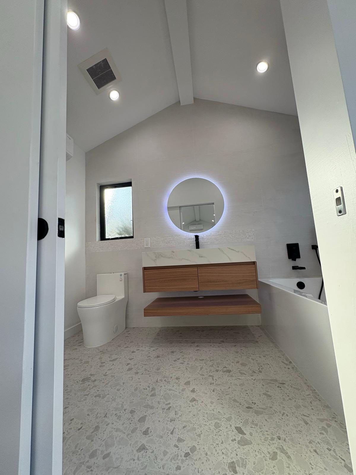 Modern white bathroom with wood vanity, round mirror, and terrazzo floor.
