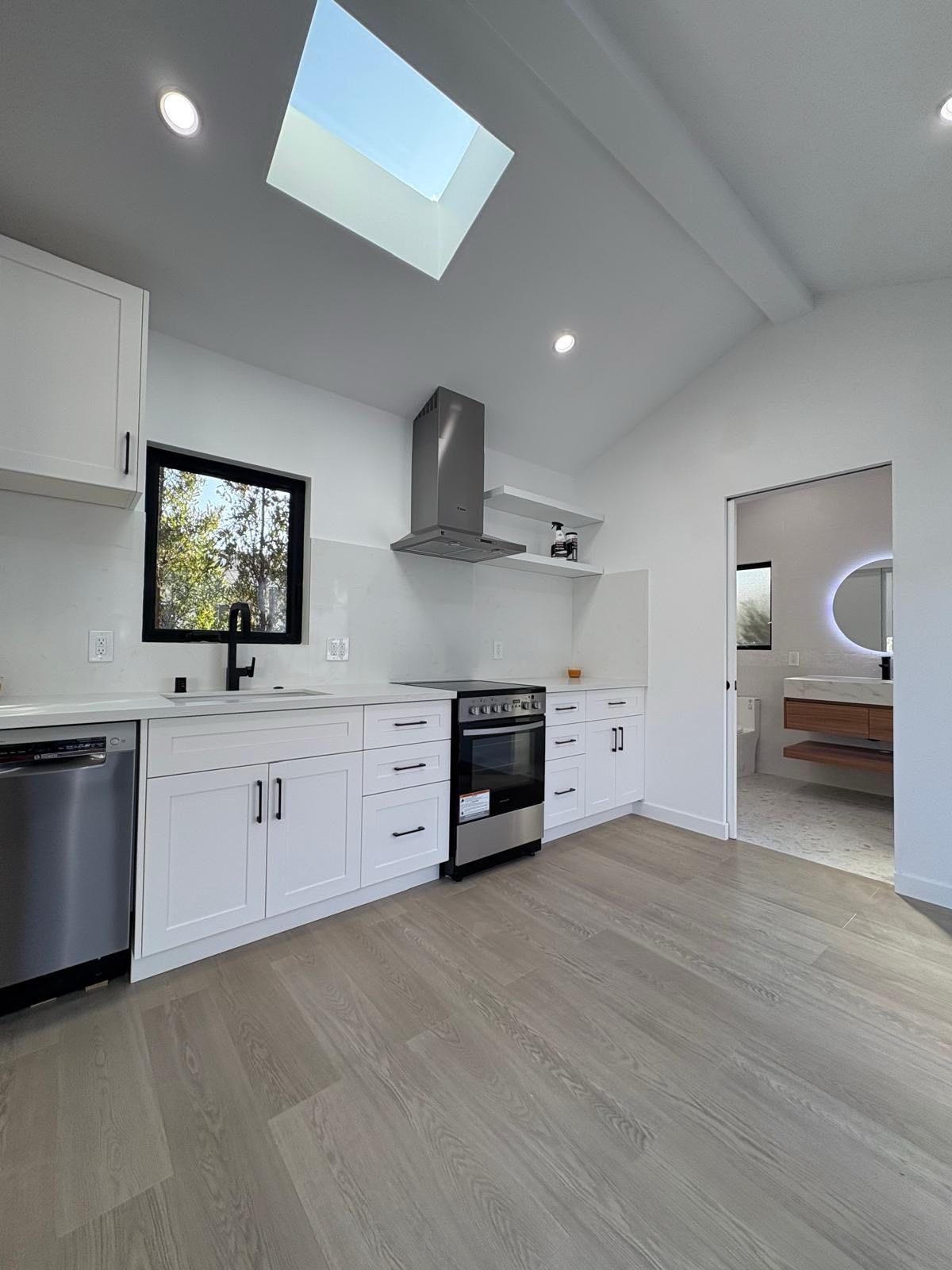 Modern white kitchen with stainless steel appliances, skylight, and doorway to a bathroom.