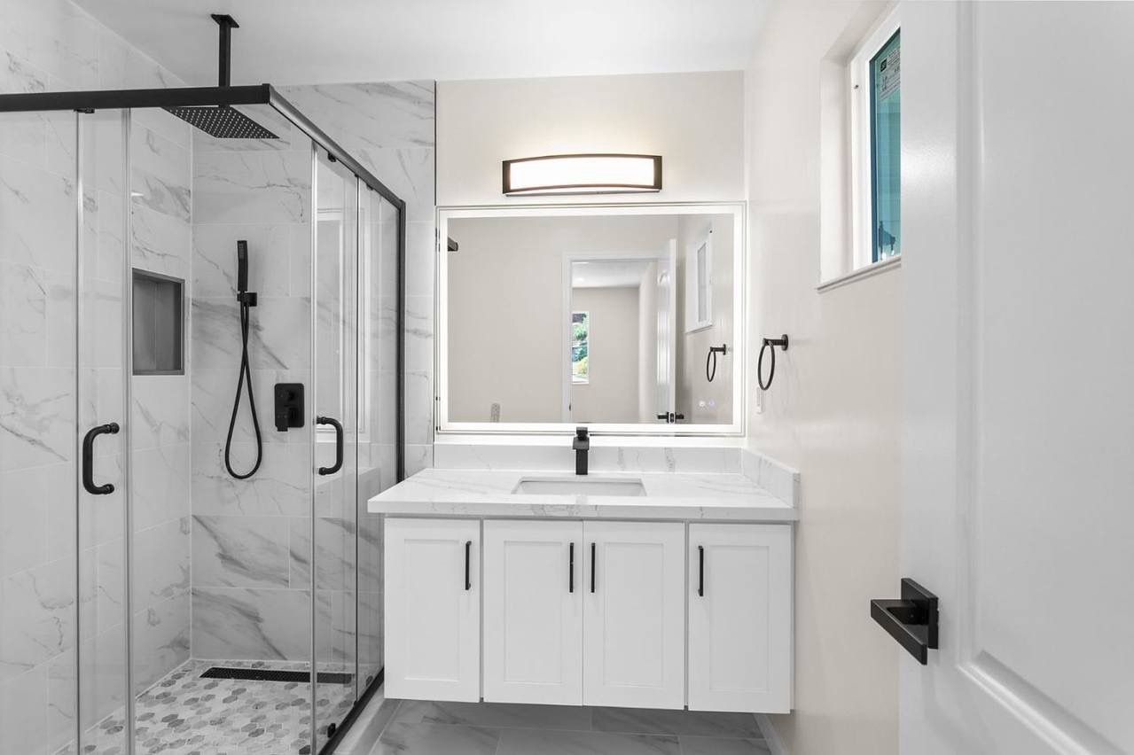 Modern white bathroom with marble shower, vanity, black fixtures, and a window.