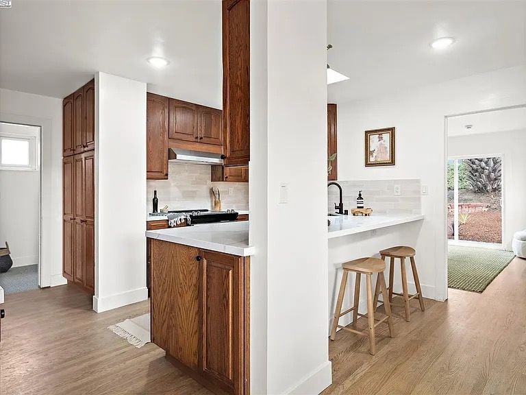 Kitchen with wooden cabinets, white countertops, and bar stools. Bright, open space with access to another room.