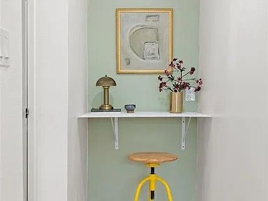 Small alcove desk with sage green wall, art, lamp, vase, and yellow stool.