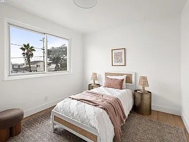 Bedroom with a white bed, pink accents, two brass side tables, and a window with a palm tree view.