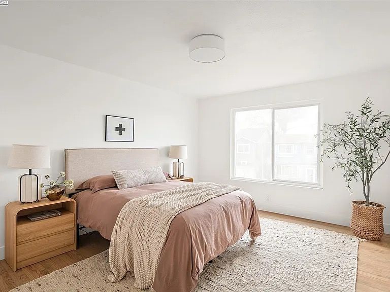 Bedroom with a bed, nightstands, rug, window, and plant. White walls and wood flooring.