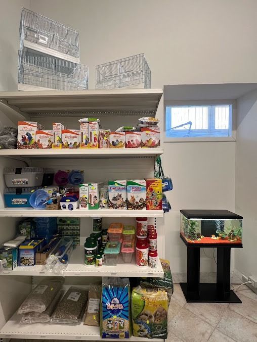 In una piccola stanza, si trovano scaffali pieni di articoli e cibo per animali domestici, con un acquario su un supporto vicino a una finestra.