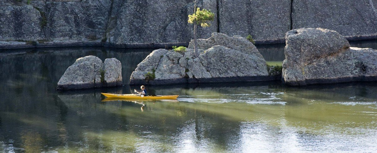 Yellow kayak gliding on a calm river beside large gray boulders and a dark stone wall