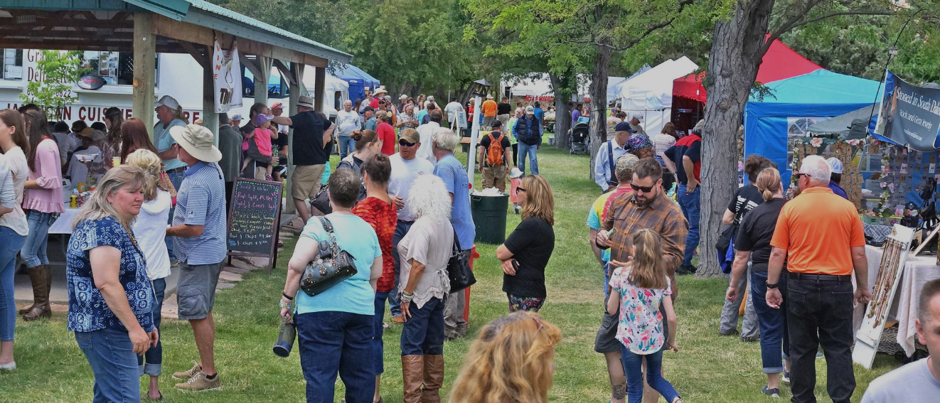 Crowd gathered at an outdoor market with tents, trees, and a small pavilion on a sunny day
