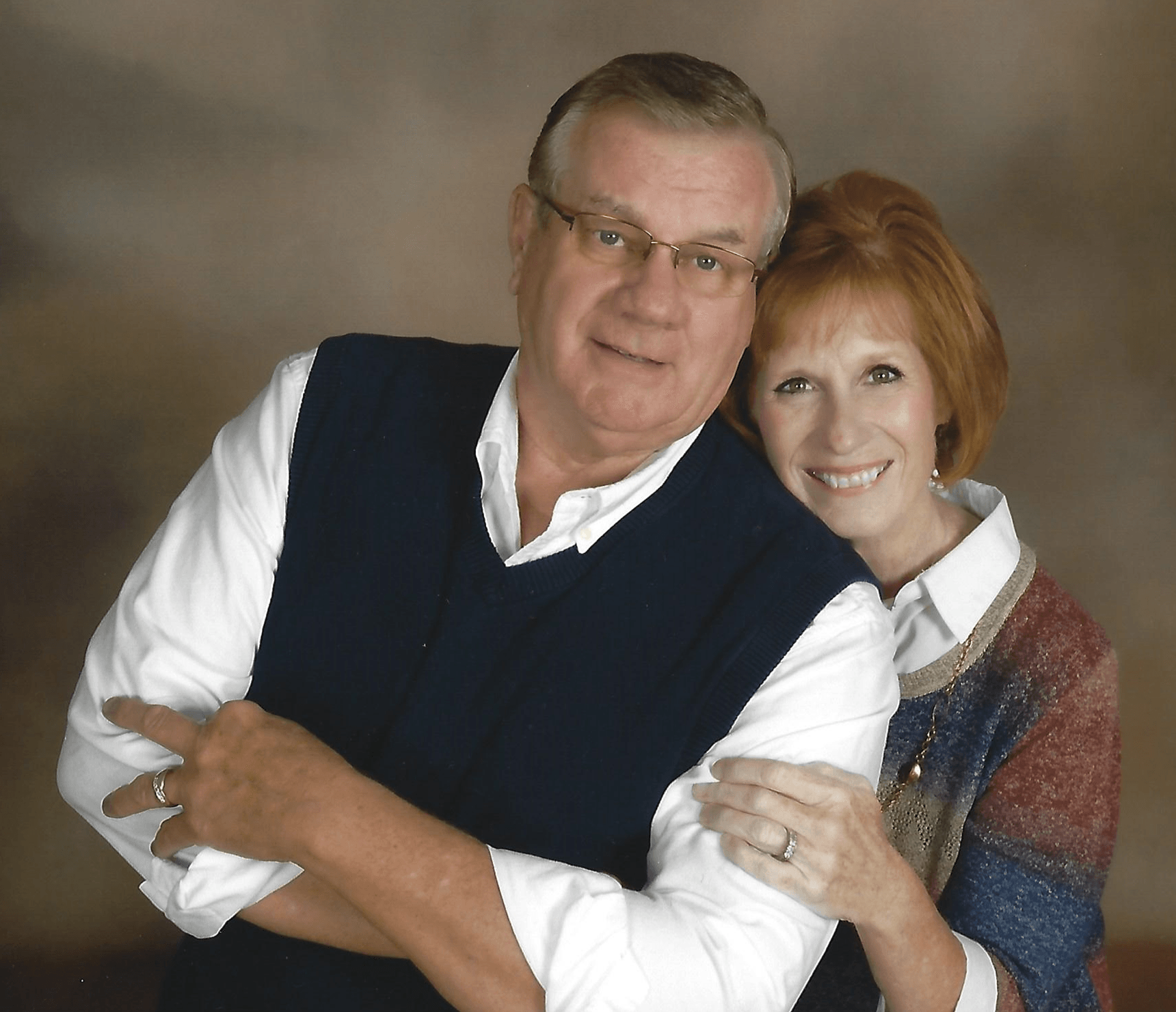 Smiling couple posing closely, man in dark vest and white shirt, woman in patterned sweater.