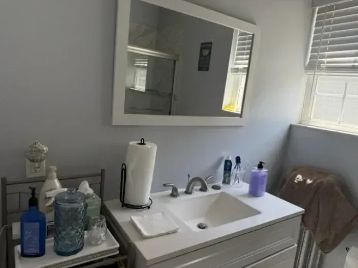Bathroom vanity with sink, mirror, and toiletries beside a window with blinds