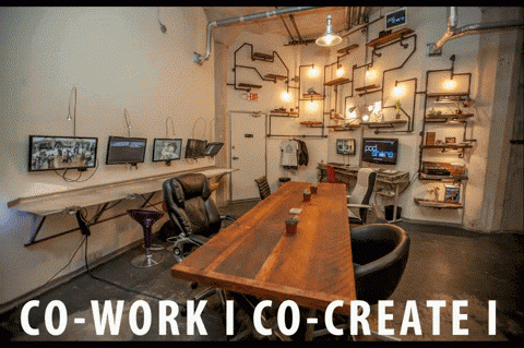 Co-working space with long wooden table, screens, industrial lighting, and exposed pipes.
